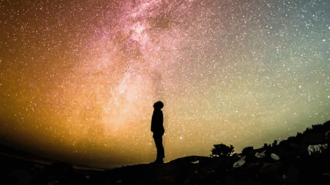 Silhouette of a person standing on a hillside, looking up at the Milky Way in a pink, orange and teal night sky