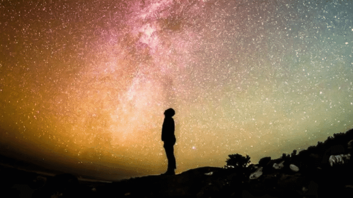 Silhouette of a person standing on a hillside, looking up at the Milky Way in a pink, orange and teal night sky
